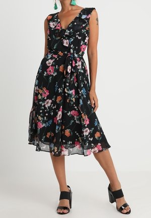 Kvinde iført en sort midi-kjole uden ærmer med farverigt blomsterprint, flæsekant ved halsen, bindebånd i taljen og sorte blokhæl-sandaler med åbne tæer.