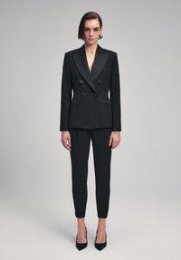 Zwarte dubbelgeknoopte blazer met satijnen revers, gecombineerd met op maat gemaakte zwarte broeken en puntige zwarte hakken, met minimale details.