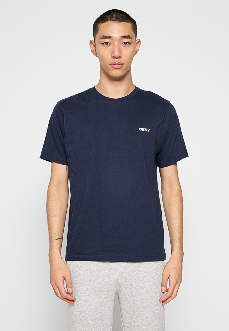 Camiseta de algodón azul marino con cuello redondo y mangas cortas, que presenta un pequeño logo blanco de DKNY en el lado izquierdo del pecho. El modelo lleva pantalones grises.