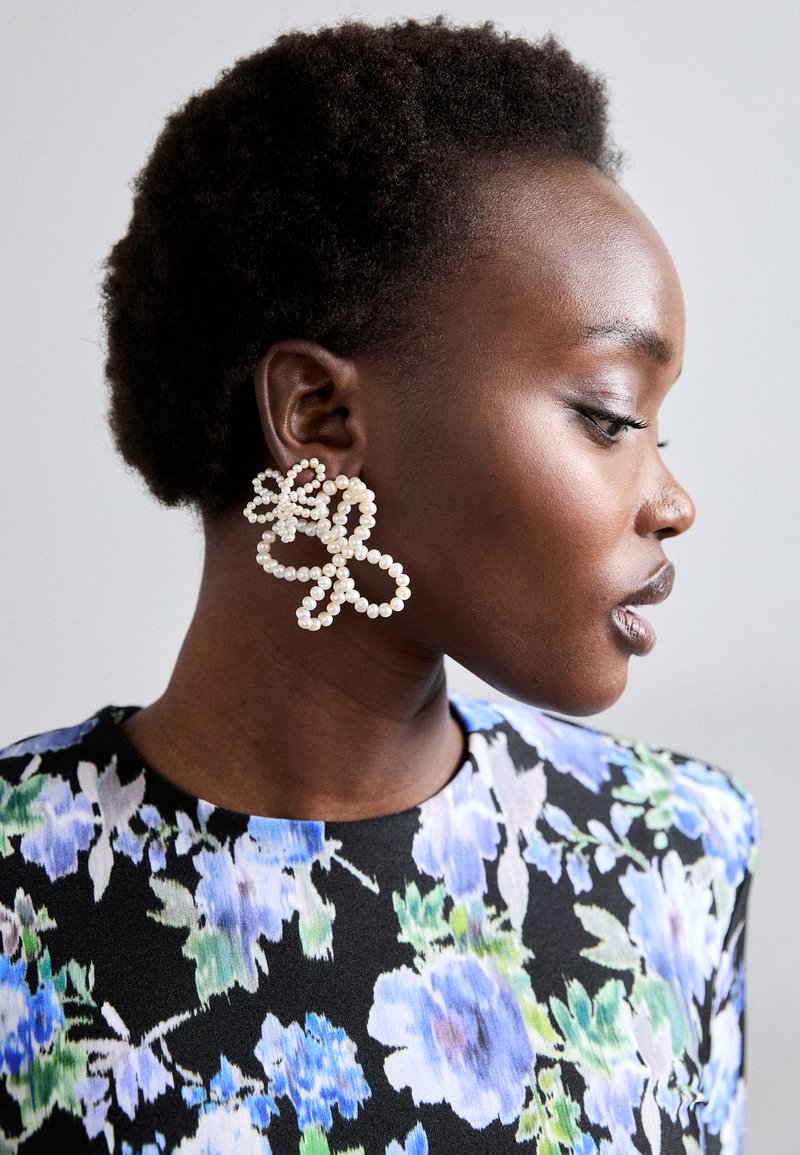 Ασημένια σκουλαρίκια με μαργαρίτες σε σχήμα λουλουδιού με υφή. Φοριούνται από ένα μοντέλο, συνδυασμένα με ένα πολυχρωμο μπλουζάκι με λουλουδάτο σχέδιο.