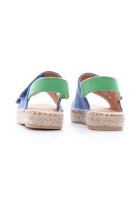 Espadrilles en toile bleue avec des bandes élastiques vertes et des semelles en corde de jute. La vue arrière met en avant la semelle plate et le talon en forme ronde.