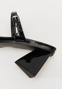 Fekete lakk bőrszíj, lapos talppal, állítható pánttal és geometrikus magas sarokkal. Simább textúra és elegáns dizájn.