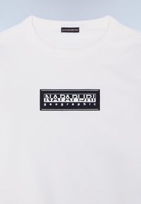 Camiseta blanca de manga corta con un logotipo rectangular negro que dice "NAPAPIJRI geographic" centrado en el pecho, con cuello redondo.