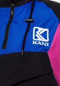 Gros plan d'une veste bleue, noire et rose avec fermeture éclair, cordon noir et logo Kani blanc brodé sur la poitrine.
