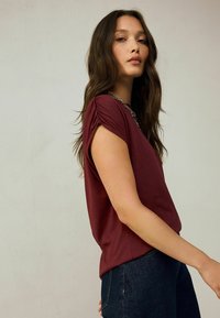 Burgundy short-sleeve top with gathered shoulder details, made of soft fabric, paired with dark blue denim jeans.