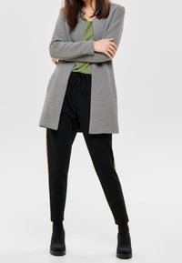 Grauer, lang geschnittene Strickjacke über einem grünen Oberteil, kombiniert mit schwarzen Jogginghosen mit einer Seitenstreife und schwarzen Stiefeletten. Glatte Texturen, lässiges Design.