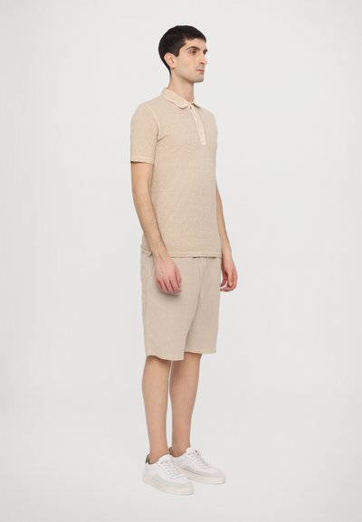 Camisa polo beige con cuello y mangas cortas, combinada con pantalones cortos beige claros y zapatillas blancas, exhibida sobre un fondo liso.
