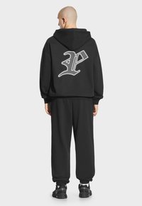 Czarny kaptur z dużym białym logo na plecach, luźny krój, zestawiony z dopasowanymi czarnymi spodniami dresowymi i czarnymi butami sportowymi.