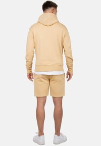 Felpa beige con cappuccio regolabile, polsini a coste e tasca a marsupio, abbinata a pantaloni corti coordinati e sneakers bianche. Texture liscia.