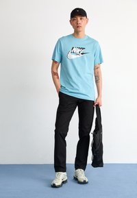 Light blue T-shirt with a black Nike logo, black pants, and light-colored athletic shoes. Black cap and a black bag in hand.