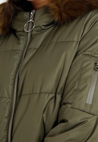 Olivgrüne Pufferjacke mit einem hohen Kragen, Reißverschluss und abnehmbarem Fellkapuze. Verfügt über Seitentaschen und eine texturierte Oberfläche.
