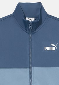 Puma jaku ar rāvējslēdzēju tumši zilā un gaiši zilā krāsā. Iespējamais augsts apkakle un balta Puma logo uz krūtīm. Gluda auduma tekstūra.