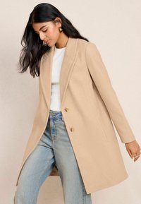 Beige wollen jas met een reverskraag, een enkelvoudige sluiting en twee knopen aan de voorkant. Gecombineerd met lichtblauwe high-waisted jeans.