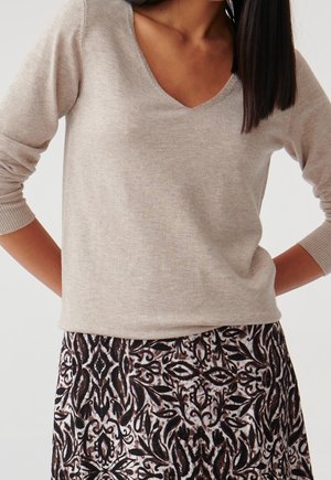 Jasnobeżowy sweter z dekoltem w serek i długimi rękawami, wykonany z miękkiego materiału. Do tego dopasowana spódnica w wzory z ciemnymi, płynnymi motywami w ciepłych, ziemistych tonacjach.