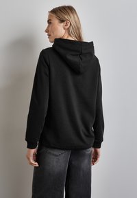 Sudadera con capucha negra hecha de un tejido suave, con un bolsillo tipo canguro y puños acanalados. Llevada con pantalones holgados de color gris oscuro.