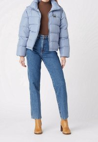 Chaqueta acolchada azul claro con un cuello alto, cierre de cremallera y un logo bordado, combinada con vaqueros de tiro alto azules y botas de tacón color beige.