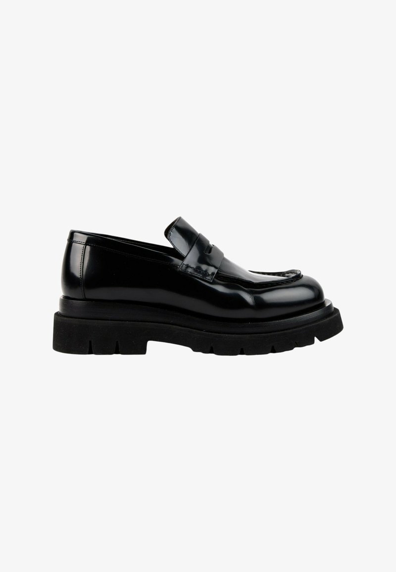 Mocassin en cuir noir brillant avec une semelle noire épaisse et épaisse et une bride sur le dessus, montré de profil sur un fond blanc.