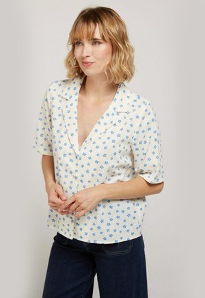 Femme aux cheveux blonds ondulés portant un chemisier blanc à manches courtes avec un motif floral bleu et un jean bleu marine, debout devant un fond uni.