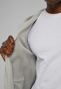 Grå lynlås hoodie med en blød tekstur, delvist åbnet, som afslører en hvid T-shirt under. Bløde stoffolder og synsdetaljer synlige.