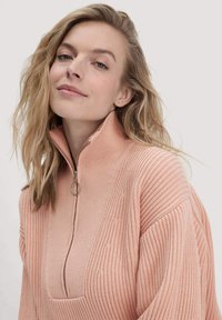 Hellrosa Strickpullover mit hohem Kragen, halbem Reißverschluss, geripptem Muster und weiten Ärmeln, der subtile vertikale Muster aufweist.