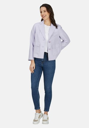 Lachende vrouw staand met hand achter de rug, draagt een lichtpaarse blazer, wit overhemd, blauwe spijkerbroek en witte sneakers op een witte achtergrond.