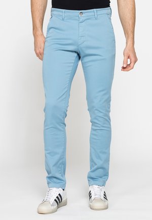 Uomo che indossa pantaloni azzurri slim fit e sneakers bianche con strisce nere, in piedi davanti a uno sfondo semplice.