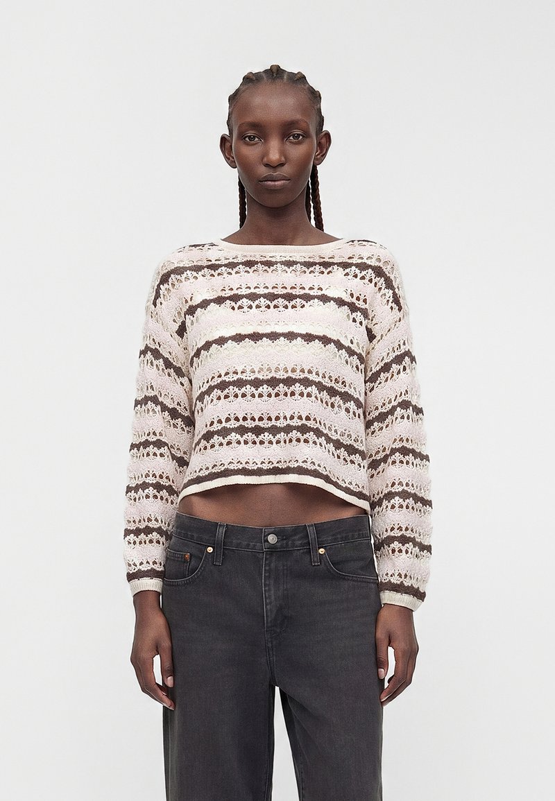 Femme portant un pull tricoté court à manches longues rayé beige et marron, associé à un jean noir taille haute, debout sur fond blanc.