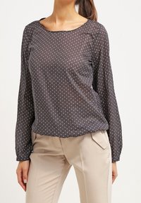 Blusa nera trasparente con pois bianchi, scollo tondo, maniche lunghe e dettagli leggermente arricciati sulle spalle. Indossata con pantaloni beige chiaro.