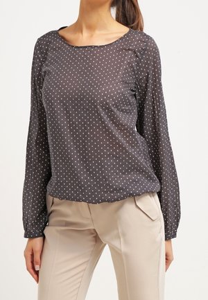 Blusa nera trasparente con pois bianchi, scollo tondo, maniche lunghe e dettagli leggermente arricciati sulle spalle. Indossata con pantaloni beige chiaro.