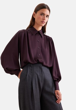 Donna che indossa una camicetta viola scuro con bottoni e maniche a sbuffo e pantaloni grigi plissettati a vita alta, posando con una mano in tasca.