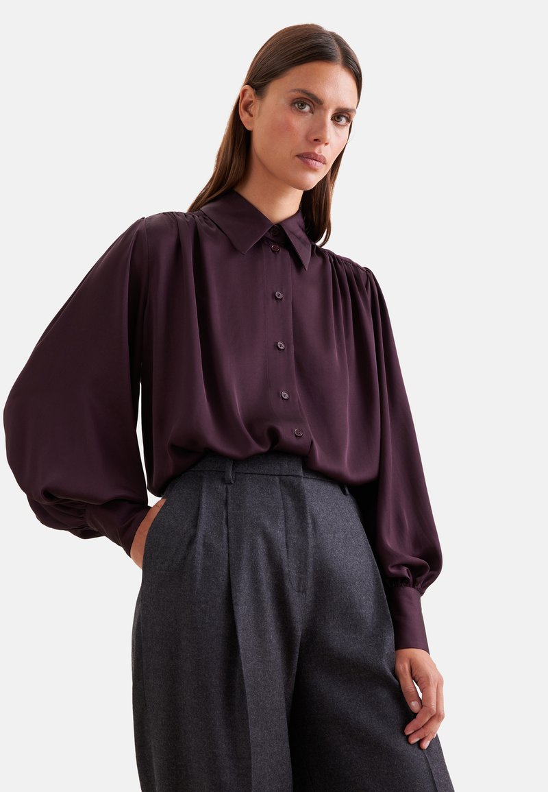 Femme portant un chemisier boutonné violet foncé à manches bouffantes et un pantalon plissé gris taille haute, posant avec une main dans la poche.