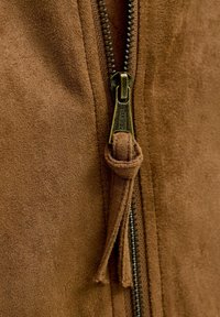 Close-up van bruin suède stof met een metalen rits en een suède trekker die in een lus is geknoopt.