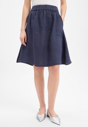 Kobieta w granatowej spódnicy do kolan z elastycznym paskiem i naszywanymi kieszeniami, w połączeniu ze srebrnymi, spiczastymi balerinami.