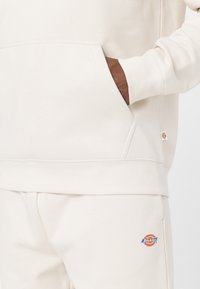 Béžová mikina s přední kapsou a žebrovanými manžetami, doplněná o odpovídající kalhoty s barevným logem Dickies. Jemný bavlněný materiál.