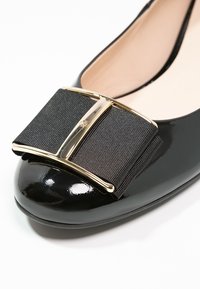 Chaussure plate en cuir verni noir avec grand nœud en tissu noir et accent en métal doré à l'avant.