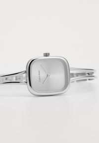 Orologio quadrato argento dal design minimalista, con un quadrante metallico liscio e un bracciale a doppia barra snodato. "Calvin Klein" è inciso.