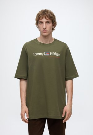 Jeune homme aux cheveux bouclés châtain clair porte un t-shirt olive Tommy Hilfiger avec logo et texte, debout devant un fond blanc uni.