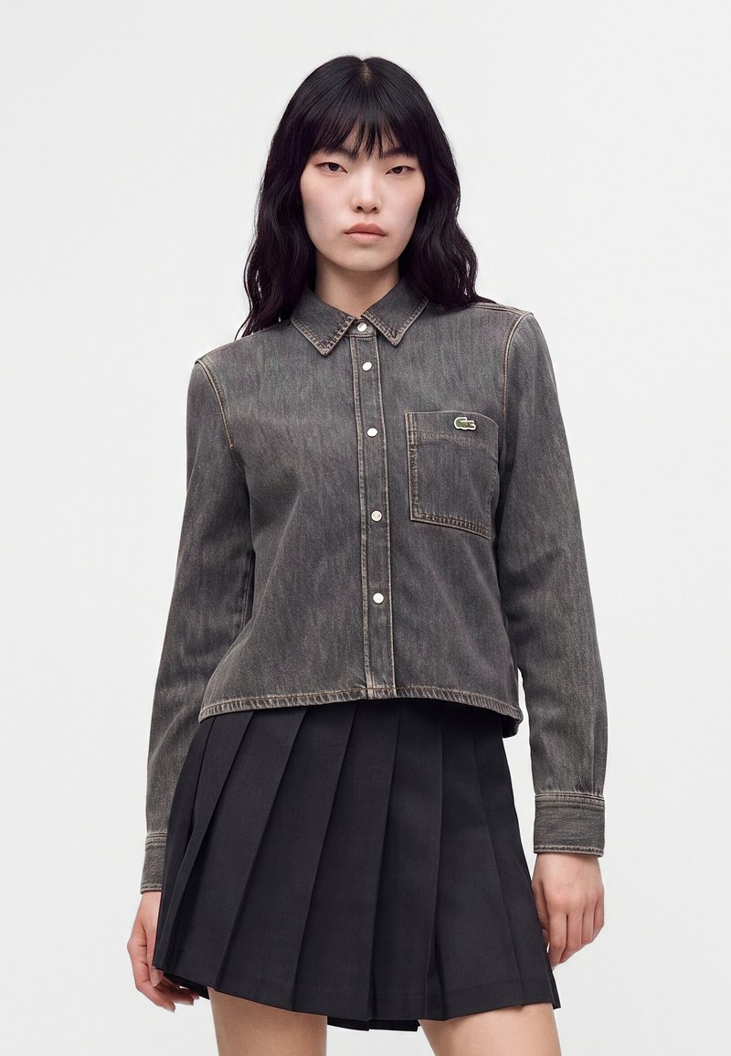 Femme aux longs cheveux noirs portant une chemise en jean noire délavée à boutons et une jupe plissée noire sur un fond clair uni.