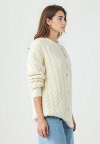 Cardigan a maglia cable beige con grandi bottoni, caratterizzato da dettagli in perle e polsini a coste. Fit oversize, abbinato a jeans azzurri.