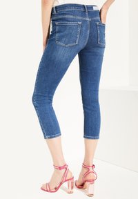 Vaqueros capri de mezclilla en azul oscuro, con un corte ajustado y longitud recortada, que presentan dos bolsillos traseros y detalles de costura sutiles. Combinados con sandalias de tacón rosas.