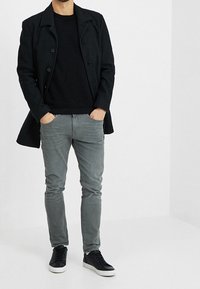 Černý vlněný kabát, texturovaný černý svetr, svetle šedé slim-fit džíny a černé tenisky s bílou podrážkou, stojící na bílém pozadí.