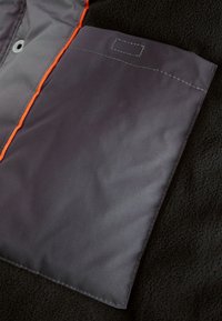 Tissu gris avec accents orange, présentant un extérieur lisse et un intérieur en polaire noire texturée ; comprend une poche plate aux bords cousus.