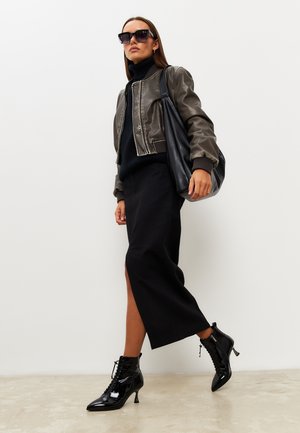 Giacca bomber in pelle marrone, dolcevita nera, gonna midi nera con spacco e stivaletti neri lucidi con punta e tacco a punta.