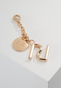 Roguldfärgad nyckelring med en stiliserad "R" och en rund tagg med ingraverad text "RIVER ISLAND." Fäst med en metallkedja och en klippkrok.