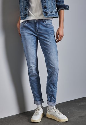 Jean en denim bleu clair avec une coupe slim et des ourlets retournés, associés à des baskets blanches. Texture lisse et détails de décoloration subtile sont visibles.