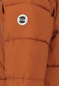 Puffet jakke i orange stof med syede sektioner. Har et rundt sort og sølv logo på ærmet og reflekterende kant langs sømmene.