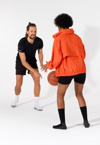 To personer, der spiller basketball. Den ene har en klar orange jakke og sorte shorts på; den anden har en sort t-shirt og shorts. Basketball i hånden.