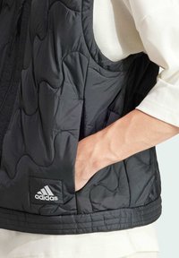 Crni kviltani prsluk s patentnim zatvaračem, s podstavljenom teksturom, zakrivljenim šavovima i Adidas logom na donjem prednjem dijelu.