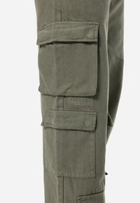 Pantaloni cargo de culoare verde măsliniu, cu multiple buzunare de depozitare, dintr-un material rezistent din bumbac și cu logo brodat pe coapsă.