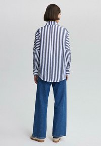 Camisa de botones a rayas en azul y blanco, con mangas largas y bajo redondeado, combinada con pantalones de mezclilla de pierna ancha y calzado ligero.